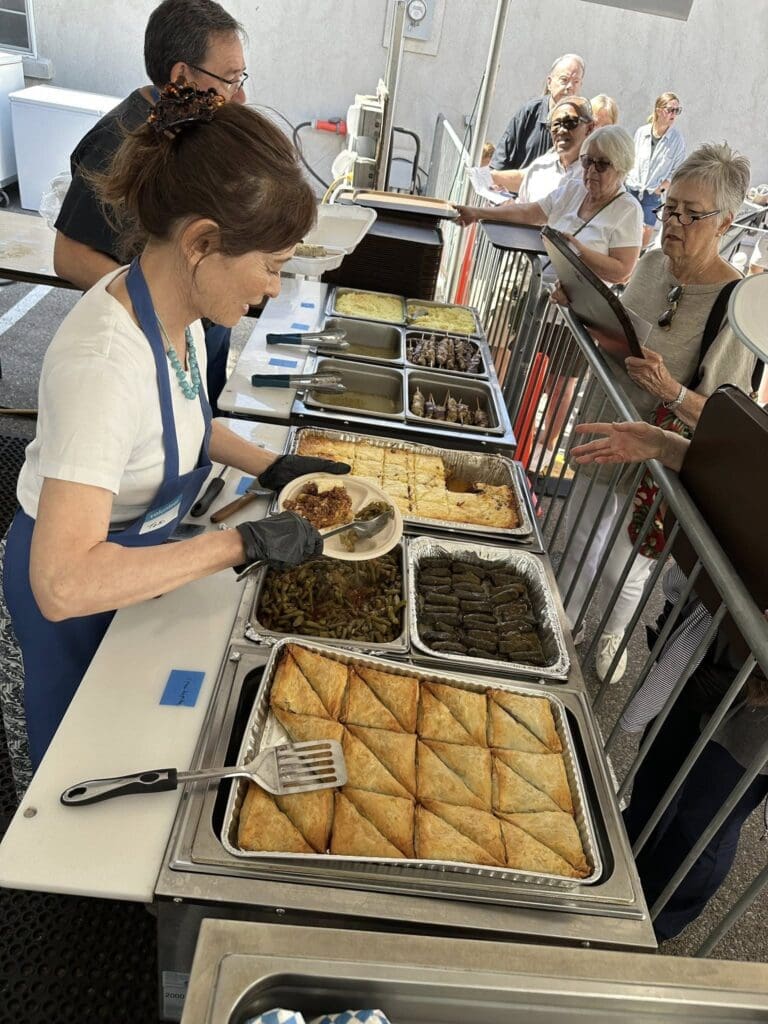 A volunteer in a blue apron and gloves serves food at an outdoor festival buffet line featuring baklava, dolmas, green beans, and other dishes, while guests wait with trays.
