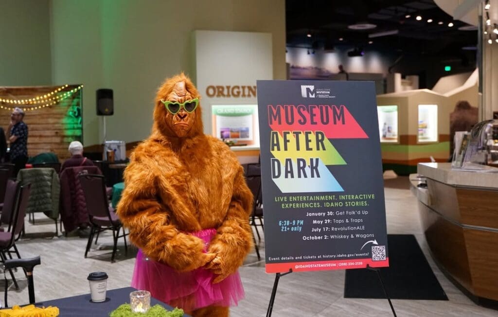 A person in a Bigfoot costume wearing green heart-shaped sunglasses and a pink tutu stands next to a 'Museum After Dark' event sign inside the Idaho State Museum.