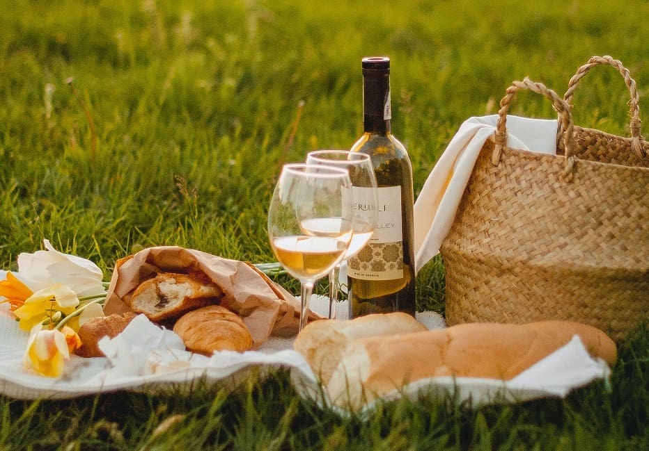 A picnic spread on a white blanket in green grass with two glasses of white wine, a bottle of wine, a baguette, pastries in paper wrapping, yellow tulips, and a woven straw bag.