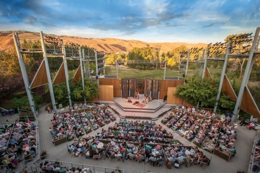 Aerial view of a packed outdoor amphitheater at dusk with a wooden stage, lush trees, and golden hills in the background.