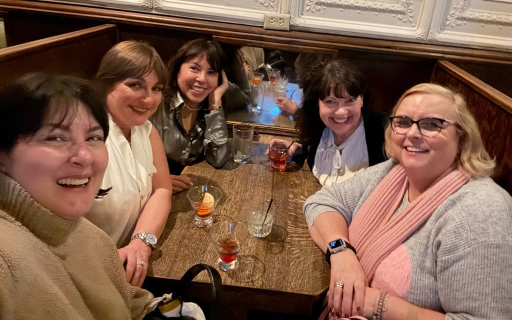 Five women smile and laugh together around a wooden table at a dimly lit bar or restaurant, with cocktails in front of them.